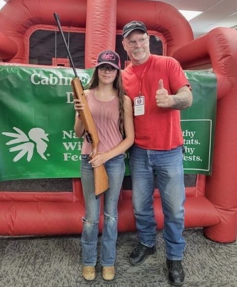 A young shooter holds a wood-stocked air rifle beside a WWCA volunteer giving a thumbs up.