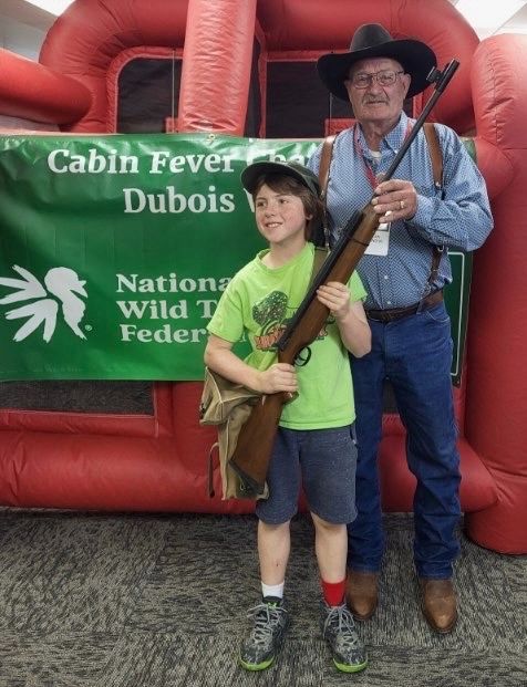 A boy in a green shirt holds a rifle beside a volunteer in a cowboy hat at the Cabin Fever NWTF banner.