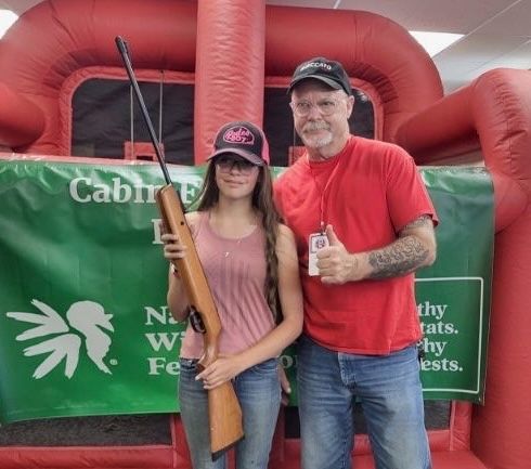 A young shooter holds a wood-stocked air rifle beside a WWCA volunteer giving a thumbs up.