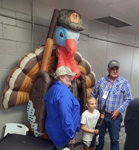 Volunteers and a young shooter beside the inflatable NWTF turkey mascot at the youth range.