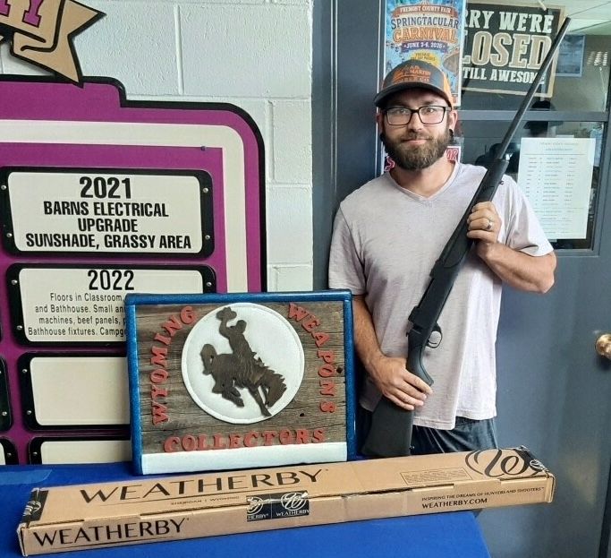 A raffle winner holds a new Weatherby rifle beside the Wyoming Weapons Collectors barnwood sign.