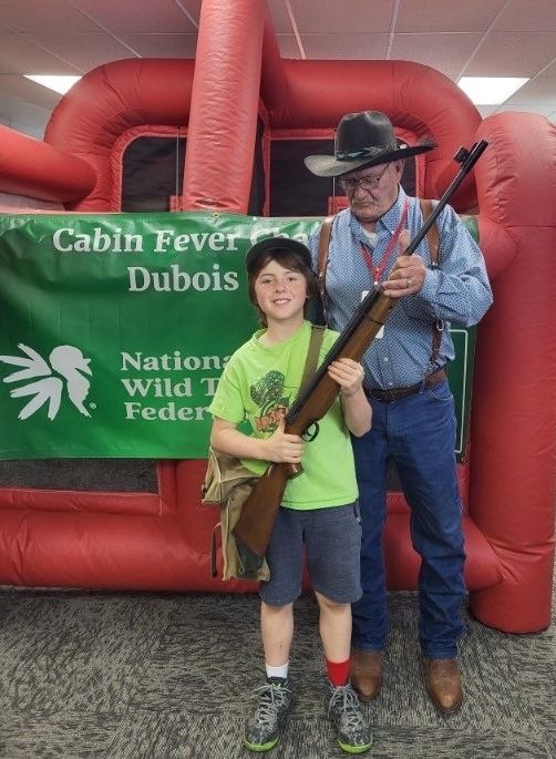 A young shooter and a cowboy-hatted volunteer pose with a rifle at the Cabin Fever NWTF banner.