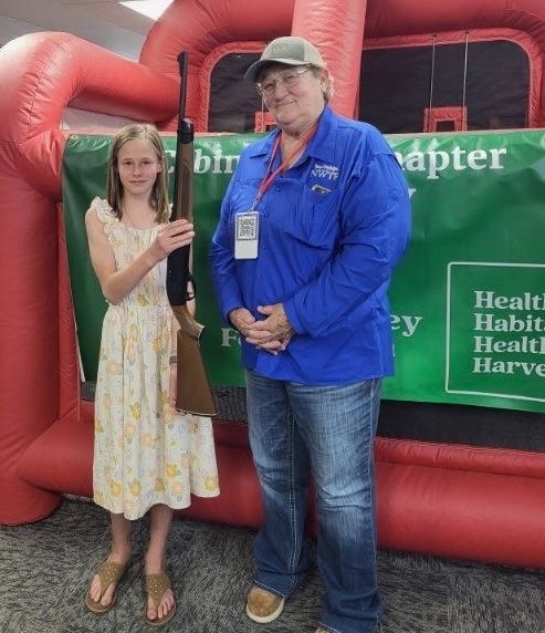 A girl in a floral dress holds an air rifle next to a National Wild Turkey Federation volunteer.