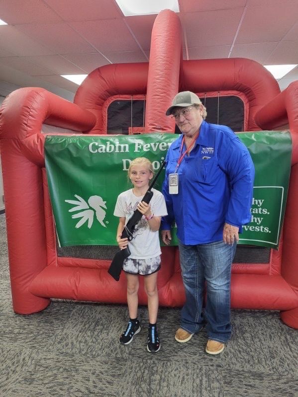 A girl holds an air rifle beside an NWTF volunteer in a blue shirt at the youth range banner.