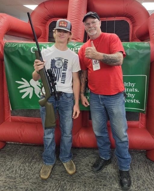 A teenager in a Lander Valley shirt holds an air rifle beside a red-shirted WWCA volunteer.